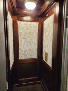 Luxury home elevator interior with dark wood panels, floral patterned wall inlays, brass control panel, and ceiling light